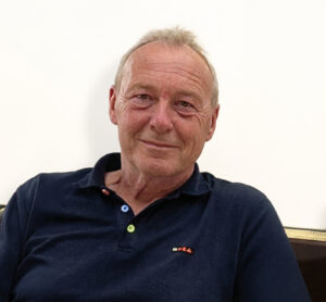 Foto von einem älteren Mann mit kurzem grauem Haar und dunkelblauem Polohemd vor weißem Hintergrund.